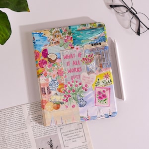 May include: A decorative journal with a collage design featuring floral arrangements, fruit, and seaside scenes. The text "WHAT IF IT ALL WORKS OUT?" is visible. A white pen and glasses are next to the journal.