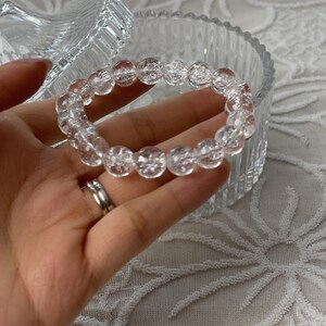 May include: A clear quartz beaded bracelet. The bracelet is made of round, transparent beads. The bracelet is held in a hand, with a clear glass container in the background. The bracelet is a piece of jewelry.