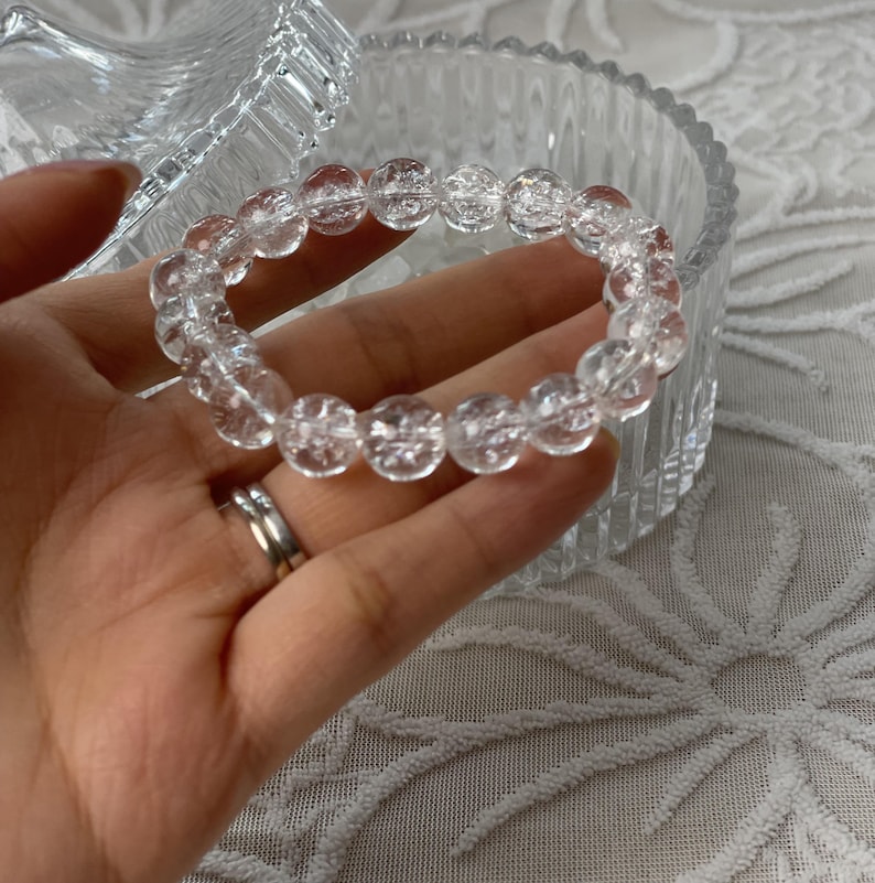 May include: A clear quartz beaded bracelet. The bracelet is made of round, transparent beads. The bracelet is held in a hand, with a clear glass container in the background. The bracelet is a piece of jewelry.