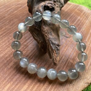 May include: A bracelet made of translucent, round, gray-green beads. The beads are strung together, forming a circular shape. The bracelet is resting on a wooden surface with a piece of driftwood.