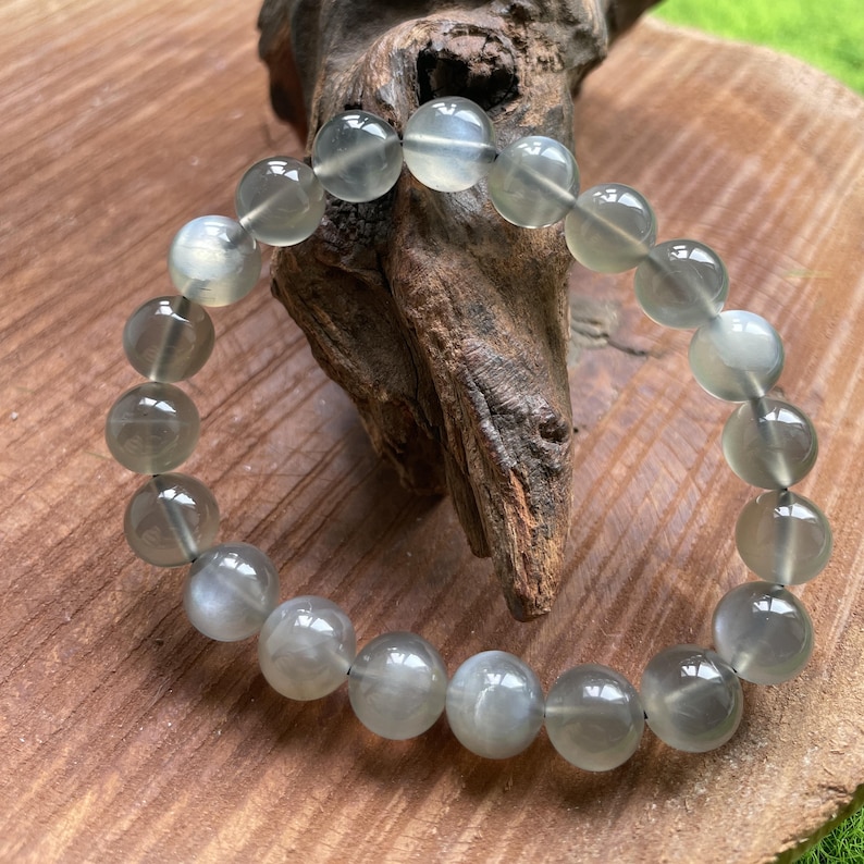 May include: A bracelet made of translucent, round, gray-green beads. The beads are strung together, forming a circular shape. The bracelet is resting on a wooden surface with a piece of driftwood.
