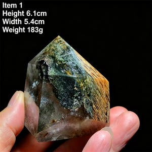 May include: A polished crystal point with a pointed top, featuring internal inclusions of dark green and golden-brown fibrous patterns. The crystal measures 6.1 cm in height, 5.4 cm in width, and weighs 183 g as indicated by the text in the image.