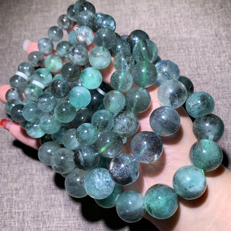 Peut inclure: Un bracelet compos&eacute; de perles rondes et polies dans des tons vari&eacute;s de vert translucide et de bleu sarcelle fonc&eacute;. Les perles pr&eacute;sentent des inclusions internes et des motifs nuageux, enfil&eacute;es sur un cordon flexible en forme de cercle.