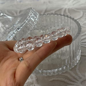 May include: Clear, faceted glass jewelry holder with a lid. A strand of clear, round beads is held in a hand. The beads are in focus, and the jewelry holder is in the background. The jewelry holder has a ribbed design.