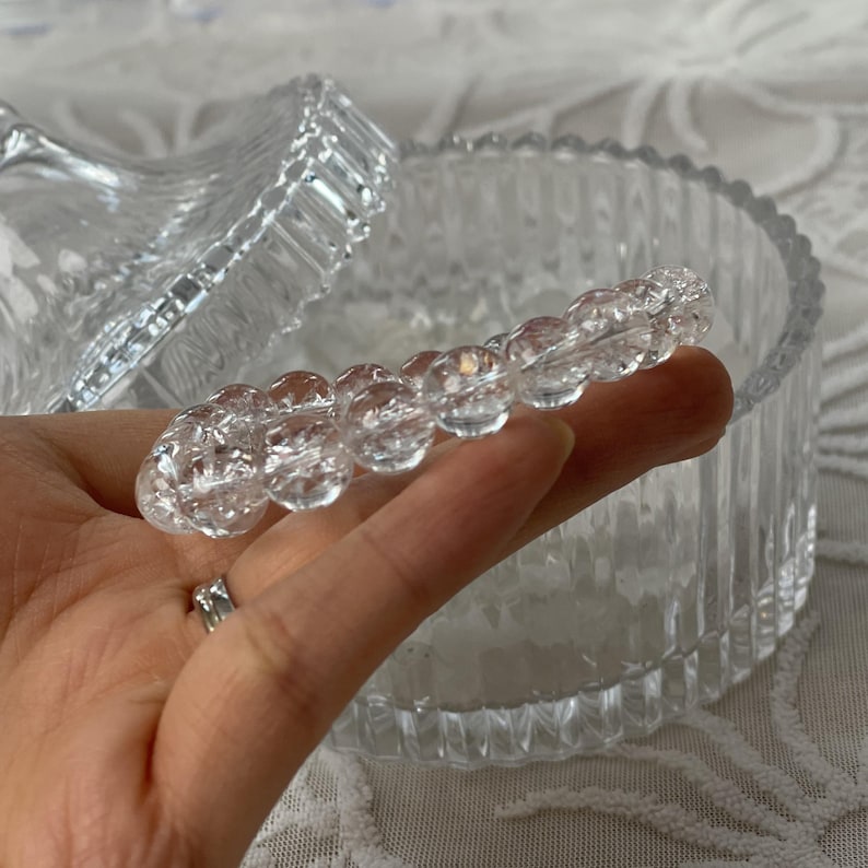 May include: Clear, faceted glass jewelry holder with a lid. A strand of clear, round beads is held in a hand. The beads are in focus, and the jewelry holder is in the background. The jewelry holder has a ribbed design.