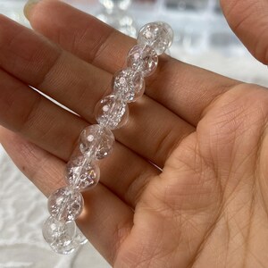May include: A clear quartz crystal bracelet. The bracelet features round, faceted beads strung together. The beads have a cracked, textured appearance, and are translucent. The bracelet is held in a hand.