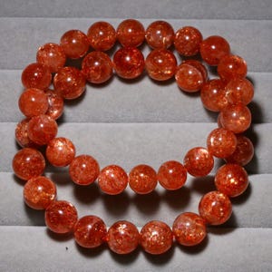 May include: A bracelet made of round, orange-red sunstone beads. The beads have a sparkling, glittery appearance. The bracelet is strung on a clear elastic cord. The beads are approximately 0.4 inches in diameter.