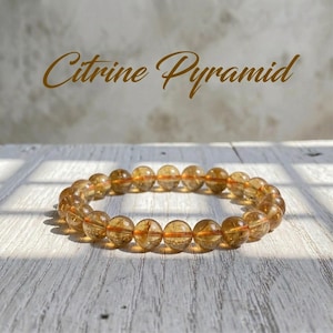 Könnte beinhalten: Ein Armband aus runden, durchscheinenden, bernsteinfarbenen Citrinperlen. Die Perlen sind aufgereiht und bilden eine kreisförmige Form. Der Text "Citrine Pyramid" steht in goldener Schrift über dem Armband.