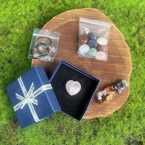 Op de afbeelding: Een verzameling kristallen en sieraden gepresenteerd op een houten oppervlak. Inclusief een blauwe geschenkdoos met een wit lint, een hartvormige rozenkwarts, een ketting en diverse kleurrijke edelstenen in kleine zakjes.