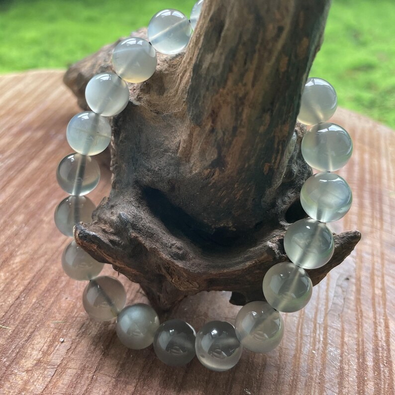 May include: A bracelet made of translucent, pale green, round beads. The beads are strung together, forming a complete circle. The bracelet is displayed on a piece of dark, weathered wood, with a blurred green background.