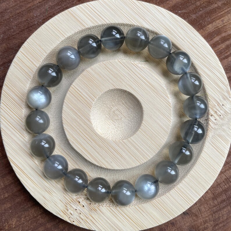 May include: A gray moonstone bead bracelet. The beads are round and have a subtle sheen. The bracelet is displayed on a light brown wooden coaster with a circular design. The beads are approximately 10mm in diameter.