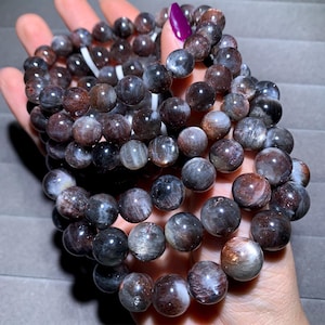 May include: A collection of beaded bracelets featuring dark, round gemstones in shades of brown, gray, and black. The beads have a polished, reflective surface, and are strung on a white elastic cord. The bracelets are held in a hand.