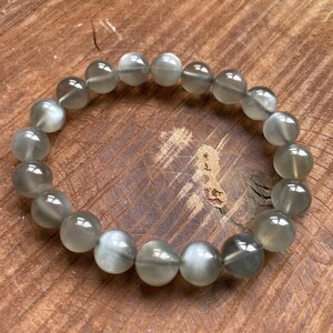 May include: A gray beaded bracelet with a mix of light and dark gray tones. The round beads are strung together, creating a circular shape. The bracelet is displayed on a wooden surface.