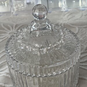 May include: A clear, decorative glass jar with a lid. The jar has a ribbed design on the sides and a beaded trim around the lid. The lid is topped with a round, clear knob. The jar is likely used for storage or display.