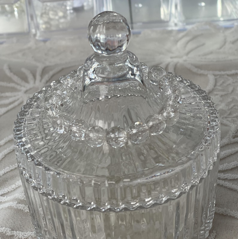 May include: A clear, decorative glass jar with a lid. The jar has a ribbed design on the sides and a beaded trim around the lid. The lid is topped with a round, clear knob. The jar is likely used for storage or display.