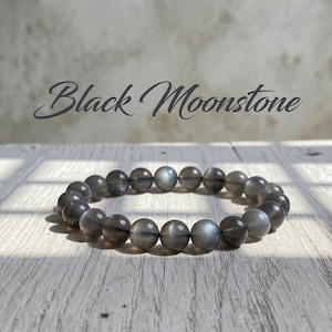 Op de afbeelding: Een armband van ronde, grijszwarte maansteen kralen. De kralen zijn aan elkaar geregen tot een cirkelvorm. De tekst "Black Moonstone" staat in cursief boven de armband.