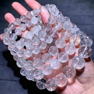 May include: A multi-strand bracelet composed of clear, spherical quartz beads. Each bead features internal fractures and natural inclusions that create a reflective, crystalline texture throughout the circular, polished stone segments strung together.