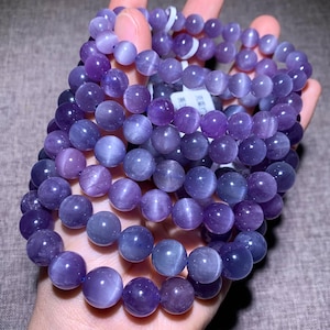 Könnte beinhalten: Mehrere Stränge lila Edelstein-Armbänder. Die Perlen sind rund und haben eine polierte, glatte Oberfläche. Die Farbe variiert von hellem Lavendel bis tiefem Violett. Die Armbänder werden in einer Hand gehalten, was die Farbvariationen und die reflektierende Qualität der Perlen zeigt.