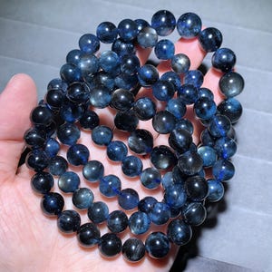 May include: A collection of dark blue gemstone beads, likely for jewelry making. The beads are round and have a polished, reflective surface. The color varies from deep navy to lighter shades of blue, creating a subtle contrast. The beads are held in a hand.