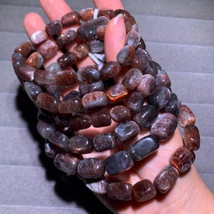 May include: A stretch bracelet composed of polished, rectangular-shaped beads in shades of deep brown, charcoal gray, and reddish-copper. The beads exhibit a natural, shimmering, and fibrous internal texture throughout the dark, multi-toned stone surface.