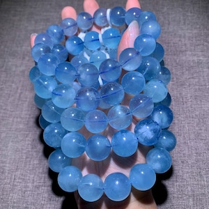 May include: A multi-strand bracelet composed of numerous round, translucent light blue beads. The beads exhibit a smooth, polished finish with subtle internal variations in clarity and color saturation, strung together on a flexible cord.