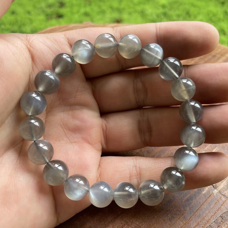 May include: A gray moonstone bead bracelet. The bracelet features round, polished beads in varying shades of gray, strung together on an elastic band. The beads have a subtle, iridescent sheen, reflecting light. The bracelet is held in a hand.