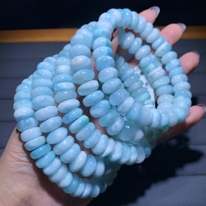 May include: Several strands of light blue gemstone bracelets. The beads are oval-shaped and have a polished finish. The bracelets are held in a hand, showcasing the color variations from light to medium blue.