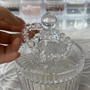 May include: A clear glass jar with a lid, featuring a decorative, clear glass bead bracelet. The jar has a ribbed design, and the bracelet is held by a hand. The background includes clear plastic storage containers.