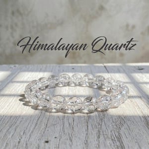 May include: A clear Himalayan Quartz bracelet with round beads. The bracelet is displayed on a light wood surface with the text "Himalayan Quartz" in a script font above it. The beads are approximately 0.5 inches in diameter.
