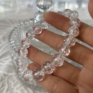 May include: A clear quartz crystal bracelet with round beads. The beads are strung together on a clear elastic cord. The bracelet is held in a hand, with a blurred background of a glass container.