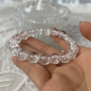 May include: A clear quartz crystal bracelet. The bracelet features multiple round, clear quartz beads strung together. The beads have internal fractures, giving them a unique, textured appearance. The bracelet is held in a hand.