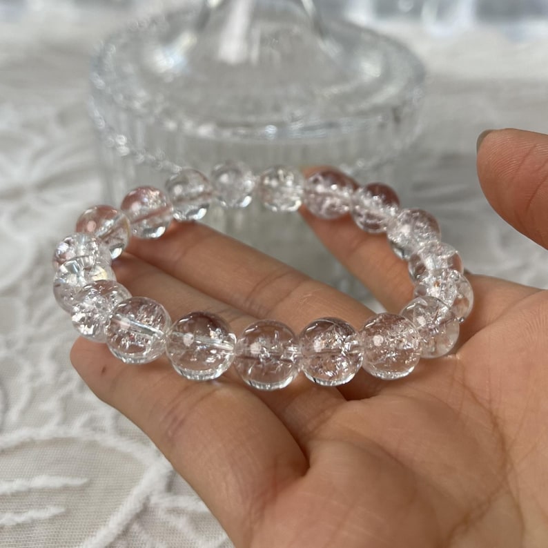 May include: A clear quartz crystal bracelet. The bracelet features multiple round, clear quartz beads strung together. The beads have internal fractures, giving them a unique, textured appearance. The bracelet is held in a hand.