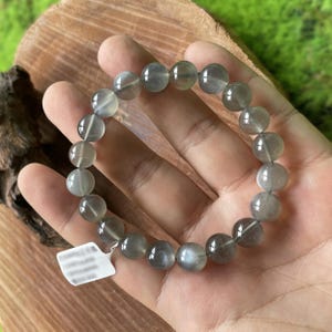 May include: A bracelet made of round, gray-toned gemstone beads. The beads are strung together, forming a complete circle. The bracelet is held in a hand, with a white tag attached. The background is a wooden surface.