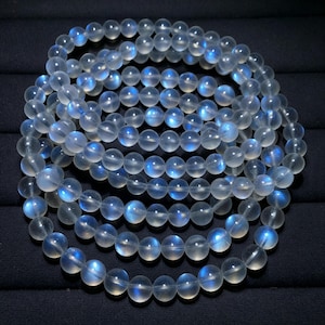 May include: A circular bracelet composed of translucent, spherical moonstone beads. Each bead features a smooth, polished surface with a distinct blue adularescence, creating a glowing effect when light hits the semi-precious stone surface.