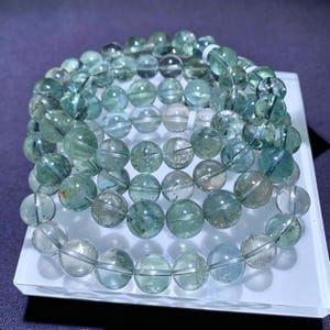May include: Several bracelets made of translucent, round, light green beads. The beads have a slightly cloudy appearance and are strung together on a clear elastic cord. The bracelets are displayed on a white surface against a purple background.