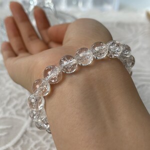 May include: A clear quartz crystal bead bracelet. The bracelet features large, round, transparent beads strung together. The beads have internal fractures and inclusions, giving them a unique appearance. The bracelet is worn on a wrist.