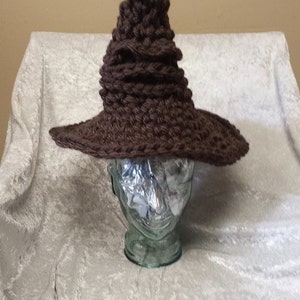Peut inclure: Un chapeau de sorcier en crochet marron avec un sommet pointu en forme de cône. Le chapeau est posé sur une tête de mannequin blanche.
