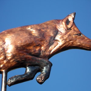 May include: A bronze-colored fox sculpture in mid-leap against a clear blue sky. The fox has detailed fur texture and is mounted on a thin metal rod. The sculpture captures the animal's dynamic movement.