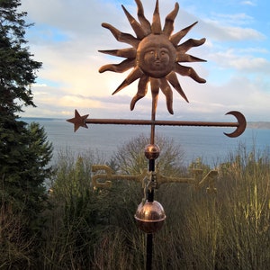 May include: A copper sun weathervane with a detailed face, star, and crescent moon. The weathervane is mounted on a black pole, with a copper sphere and directional indicators. The background features a view of the sea and sky.