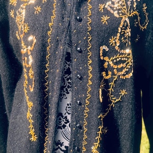 Puede incluir: Cárdigan negro con bordados dorados y adornos de lentejuelas. El suéter presenta un cuello redondo, mangas largas y cierre de botones. Los bordados y lentejuelas doradas forman un diseño floral.