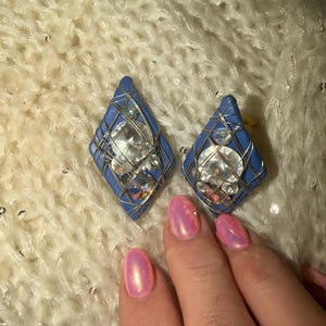 May include: A pair of blue and silver diamond-shaped earrings. Each earring features a large, clear gemstone encased in a silver wire cage, set against a blue background. The earrings are displayed on a textured, off-white surface.