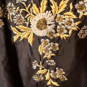 May include: Close-up of a black fabric with intricate floral embroidery. The embroidery features gold, silver, and brown thread, creating a detailed and textured design. The flowers and leaves are meticulously stitched.