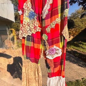 May include: Brightly colored plaid pants with lace and floral embellishments. The pants feature a red, orange, yellow, and black plaid pattern, with patches of cream and white lace, and embroidered floral details. The pants are hanging outside.