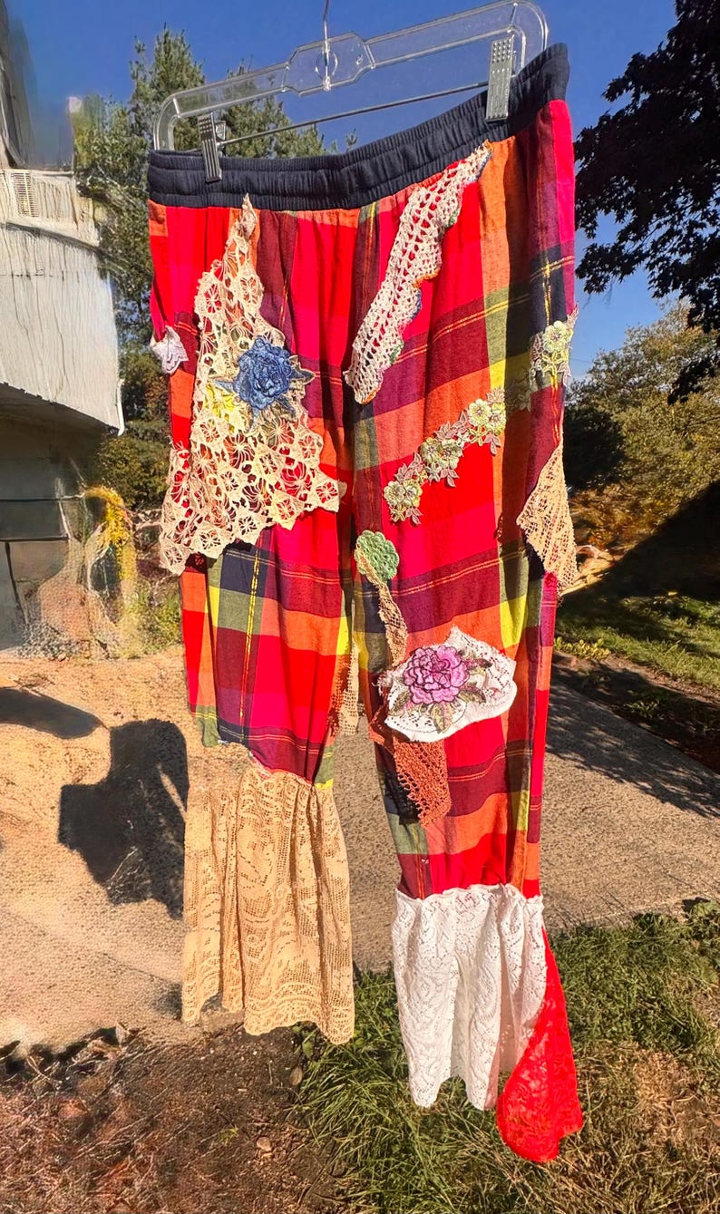 May include: Brightly colored plaid pants with lace and floral embellishments. The pants feature a red, orange, yellow, and black plaid pattern, with patches of cream and white lace, and embroidered floral details. The pants are hanging outside.