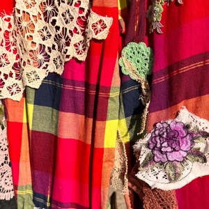 May include: A vibrant, patchwork textile featuring a plaid pattern in red, pink, yellow, and blue. The fabric is adorned with various crochet and lace appliqu&eacute;s, including floral and geometric designs. The bottom edge has a lace trim.