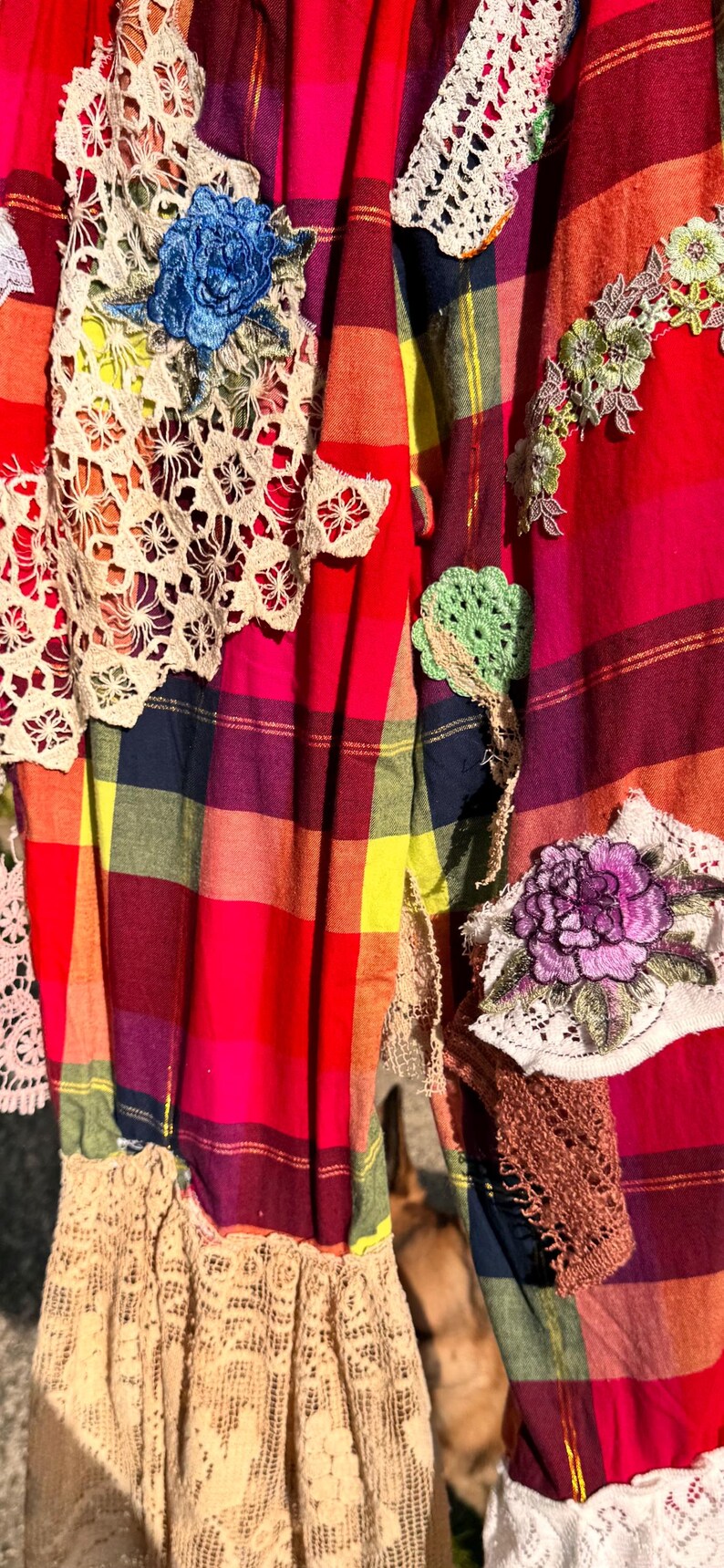 May include: A vibrant, patchwork textile featuring a plaid pattern in red, pink, yellow, and blue. The fabric is adorned with various crochet and lace appliqu&eacute;s, including floral and geometric designs. The bottom edge has a lace trim.