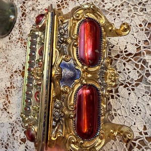 May include: Ornate gold-colored metal frame with two large, red, oval-shaped jewels. The frame has intricate detailing and a blue accent. The object appears to be a decorative piece or a small box.