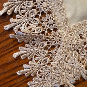 May include: Close-up of a beige lace trim with intricate floral and leaf patterns. The lace is sewn onto a cream-colored fabric, creating a delicate and elegant design. The trim is placed on a wooden surface.