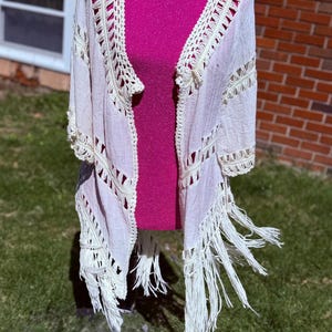 May include: A cream-colored, open-front kimono-style cardigan with crochet detailing along the edges and sleeves. The cardigan has fringe along the bottom. The garment is displayed on a pink mannequin.