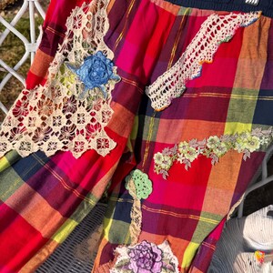May include: Colorful plaid pants with decorative lace and floral patches. The pants feature a mix of red, pink, yellow, and blue plaid, with various lace and embroidered floral accents. The waistband is a dark blue.